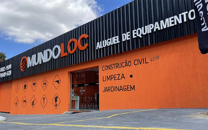 Fachada da loja MundoLoc com muro alaranjado e placa preta com estacionamento na porta.
